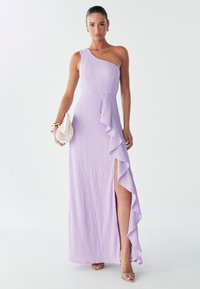 Maximekko, jossa on yksiolkaiminen laventelinvärinen muotoilu, teksturoitu kangas, sivuröyhelöyksityiskohtia ja reittä korkealle ulottuva halkio. Yhdistetty pienen valkoisen laukun ja hihnallisten sandaalien kanssa.