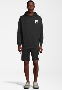 Czarna bluza z kapturem z przednią kieszenią i białym, teksturowanym logo, zestawiona z dopasowanymi szortami. Nosi się ją z szarymi butami sportowymi. Prosty, casualowy design.