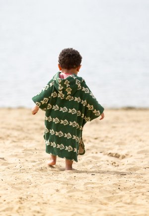 Toddler walking barefoot on sandy beach wearing green hooded towel with white fish pattern, facing away from camera.