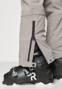 Hallid suusapüksid on pahkluu juures tõmblukuga avatavad, paljastades musta suusasaapa, millel on metallist detailid ja valge bränding.