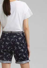 Námořnické květinové šortky s bílými vzory listů, srolovanými manžetami a plochým pasem, v kombinaci s jednoduchou bílou košilí s krátkým rukávem.