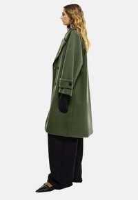 Olivgrüner Wollmantel mit oversize Passform, großen Revers, doppelter Knopfleiste, zwei Knöpfen und seitlichen Taschen. Schwarze Handschuhe und Hose vervollständigen den Look.
