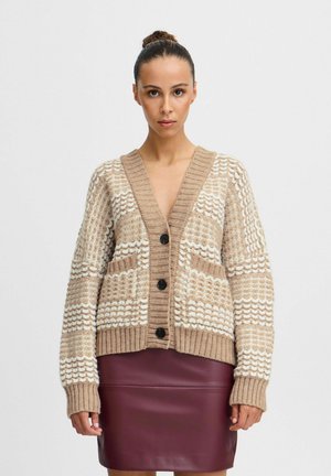 Gilet beige avec des motifs blancs texturés, des poignets côtelés et un décolleté en V, orné de boutons noirs et de deux poches avant, associé à une jupe bordeaux.