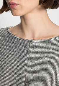 Harmaa ja valkoinen raidoitettu paita, jossa on leveä, pyöreä pääntie. Kankaan pinta näyttää pehmeältä, ja siinä on vinoraitakuosi sekä hienovaraisia ompelusaumoja.