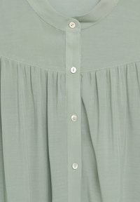 Jasnozielona bluzka z okrągłym dekoltem, zapięciem na guziki z guziczkami przypominającymi perły oraz miękką, marszczoną strukturą materiału.