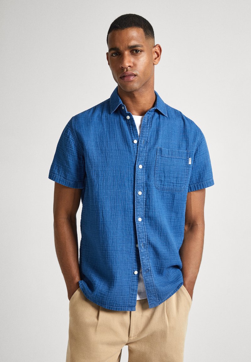 Camisa azul de botón de manga corta hecha de tela texturizada, con un bolsillo en el pecho y cuello clásico, llevada sobre una camiseta blanca.