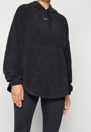 Person trägt einen lockeren schwarzen Fleece-Hoodie mit kleinem weißem Nike-Swoosh und schwarze Leggings, steht vor einem einfarbigen hellen Hintergrund.