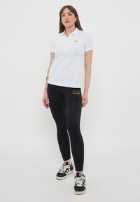 Polo bianco con un piccolo logo dorato, abbinato a leggings neri con testo dorato e sneakers nere con accenti bianchi.