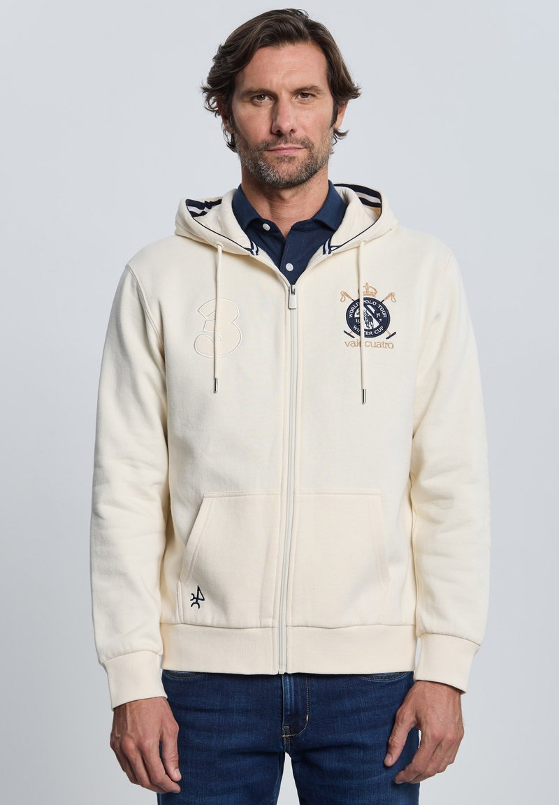 Sudadera con cremallera color crema con detalles en azul marino, con el número "3" en el pecho y detalle de logo. Hecha de material suave con bolsillos tipo canguro.