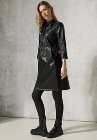 Schwarzes Lederkleid mit Kragen, Dreiviertelärmeln, seitlichen Taschen und ausgestelltem Saum. Kombiniert mit schwarzen klobigen Stiefeln und Strumpfhosen.