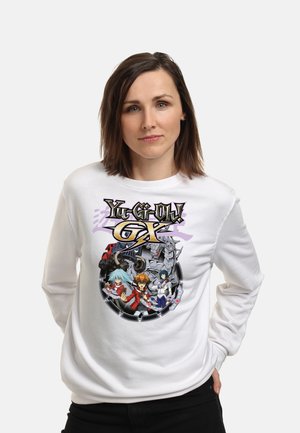 Kvinde iført en hvid sweatshirt med farverige Yu-Gi-Oh! GX anime-figurer og logo, stående med den ene hånd i lommen mod en ensfarvet baggrund.