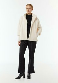 Crème-kleurige nepbontjas met een brede kraag, drie knopen en lange mouwen, gecombineerd met zwarte slim-fit broek en stijlvolle zwarte hakken.