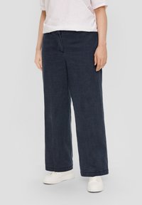 Wijduitlopende broek van donkerblauw stof, met een gladde textuur en een ontspannen pasvorm. Gecombineerd met witte sneakers voor contrast.