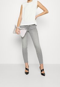 Mujer vestida con blusa blanca sin mangas, jeans ajustados gris, tacones puntiagudos negros, sosteniendo un bolso de mano plateado texturizado, de pie contra un fondo blanco.