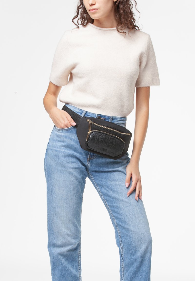 Marsupio nero con superficie testurizzata e zip dorata, indossato sopra jeans azzurri chiari e un maglione corto crema, esposto su uno sfondo bianco.