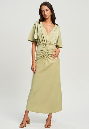Woman wearing a light green ruched midi dress with short flutter sleeves and tan strappy heeled sandals, standing against a plain background.