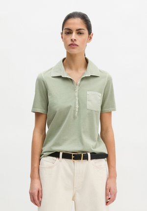 Kobieta ubrana w jasnozieloną koszulkę polo z krótkim rękawem i kieszenią na piersi, wpuszczoną w białe spodnie z czarnym paskiem, stojąca na białym tle.