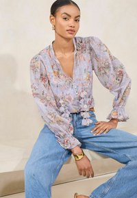 Bluză florală din material transparent în lavandă deschisă, cu mâneci tip balon, detalii cu volane și închidere cu nasturi pe față. Este asortată cu blugi blue jeans cu talie înaltă.