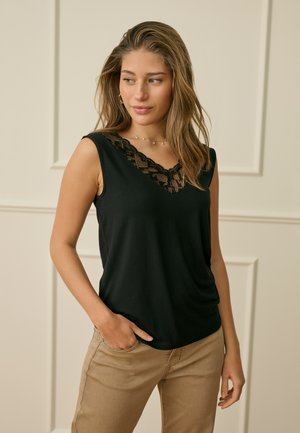 Femme aux cheveux ondulés portant un haut noir sans manches à encolure en dentelle et un pantalon beige, debout avec une main dans la poche, regardant vers la gauche.