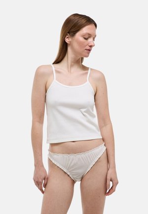 Femme portant un crop top blanc sans manches et un sous-vêtement bikini beige clair à volants, debout devant un fond clair uni.