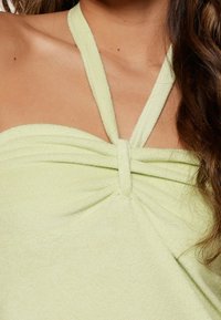 Top halter verde claro hecho de una tela suave, con un detalle de nudo torcido en el centro y tirantes delgados que se extienden sobre los hombros.