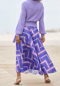 Fioletowy sweter dzianinowy z luźnymi rękawami, w połączeniu z długą, zwiewną spódnicą w geometryczny różowo-niebieski wzór. Płaskie sandały.