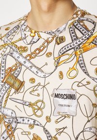 Koszulka z beżowym tłem, z nadrukiem narzędzi krawieckich, centymetrów, guzików i nożyczek w złotych i czarnych akcentach.