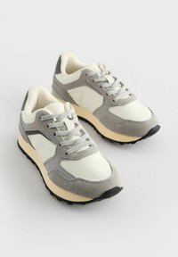 Sneaker mit einem Obermaterial aus cremefarbenem Leder und grauen Wildlederelementen, Schnürung an der Vorderseite, gepolsterter Kragen und einer strukturierten Sohle aus schwarzem Gummi.