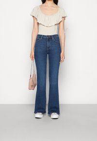 Udsvingede blå jeans med høj talje, parret med en beige top med en flæse ved halsen, og holder en lille lyserød håndtaske.