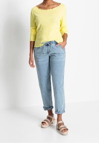 Žlutý žebrovaný top s dlouhým rukávem a vlnitými okraji, spárovaný s světlemodrými ohrnutými džíny a béžovými platformovými sandály.