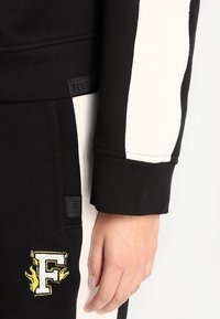 Felpa atletica nera e crema con texture a coste, logo ricamato e design dei pannelli laterali. Include un'etichetta in tessuto.