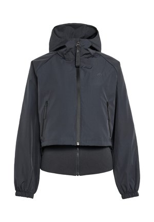 Jong persoon draagt een zwarte hoodie, losse zwarte broek, zwarte sneakers en draagt een zwarte rugzak, staat tegen een effen achtergrond.