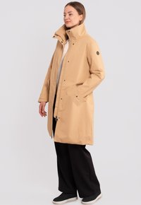 Beige vettovallintainen takki, jossa on korkea kaulus, etuosan napitus ja sivutaskut, yhdistettynä mustiin housuihin ja tennareihin. Suuressa on sileä pinta.