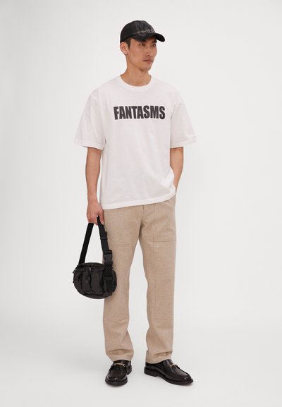 Hvit T-skjorte med svart "FANTASMS" tekst, beige bukser, subtil tekstur, svart crossover-veske, svarte sko med gull detaljer, avslappet passform.
