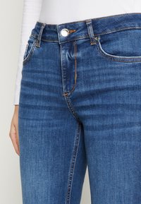 Primer plano de unos vaqueros de denim azul de corte medio con costuras naranjas y un botón plateado, llevados con una camisa blanca de manga larga visible en la cintura y el brazo.