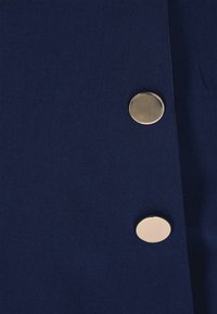 Sötétkék szövet, rajta két nagy, fényes, aranyszínű, kerek gomb függőlegesen rendezve a jobb oldalon.