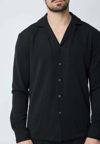 Chemise noire à manches longues en tissu texturé ; dotée d'un col camp et de cinq boutons sur le devant. Design simple et épuré avec une coupe décontractée.