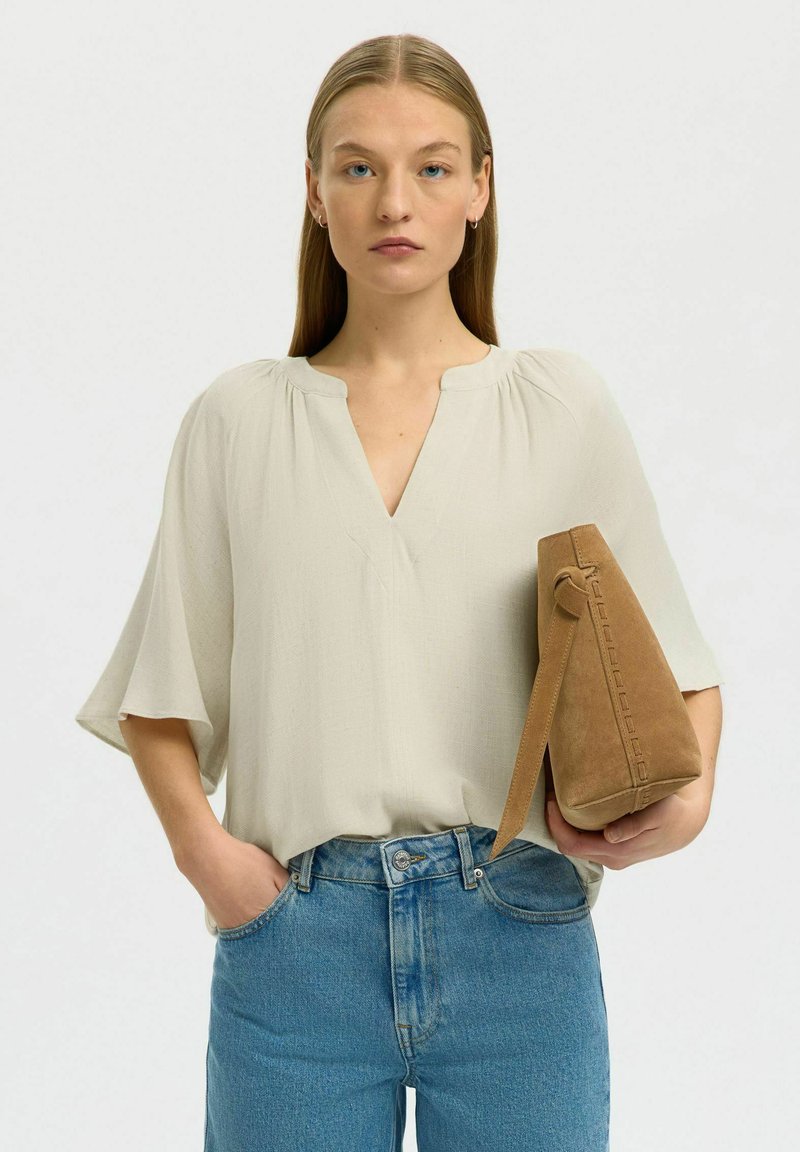 Femme aux cheveux blonds et raides portant un chemisier crème et un jean bleu, tenant un sac à main en daim marron avec une poignée supérieure.