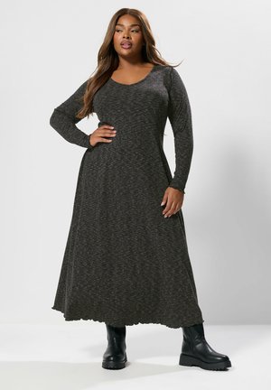 Vrouw met rondingen in een lange, donkergrijze maxi-jurk met lange mouwen en zwarte enkellaarsjes, staande met één hand op haar heup tegen een effen achtergrond.