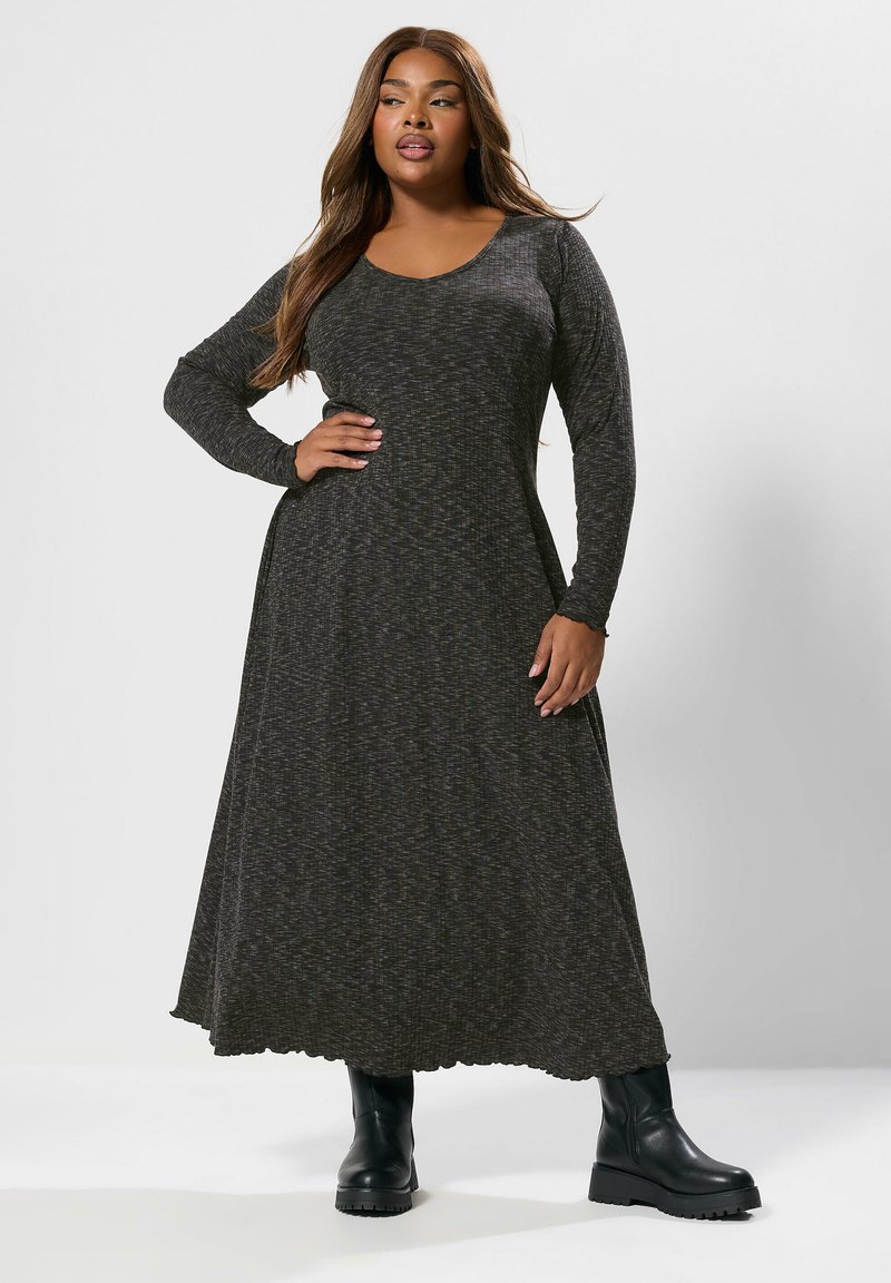 Eine kurvenreiche Frau trägt ein langärmliges, dunkelgraues Maxikleid mit schwarzen Knöchelstiefeln und steht mit einer Hand auf der Hüfte vor einem schlichten Hintergrund.
