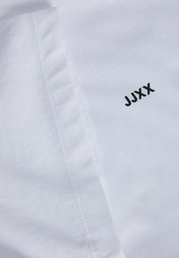 Hvidt stof med detaljerede syninger og foldet kant, med sort tekst "JJXX" trykt på overfladen.