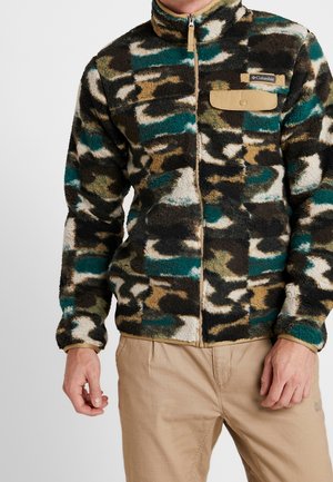Ein Mann trägt eine Fleecejacke im Camouflage-Muster in Grün, Braun, Schwarz und Weiß mit einer beigen Druckknopftasche und hellbraunem Reißverschluss, kombiniert mit beigen Hosen.