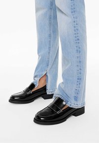 Mocassins en cuir verni noir à talons blocs, avec coutures contrastées et côtés découpés, portés avec un jean en denim bleu clair.