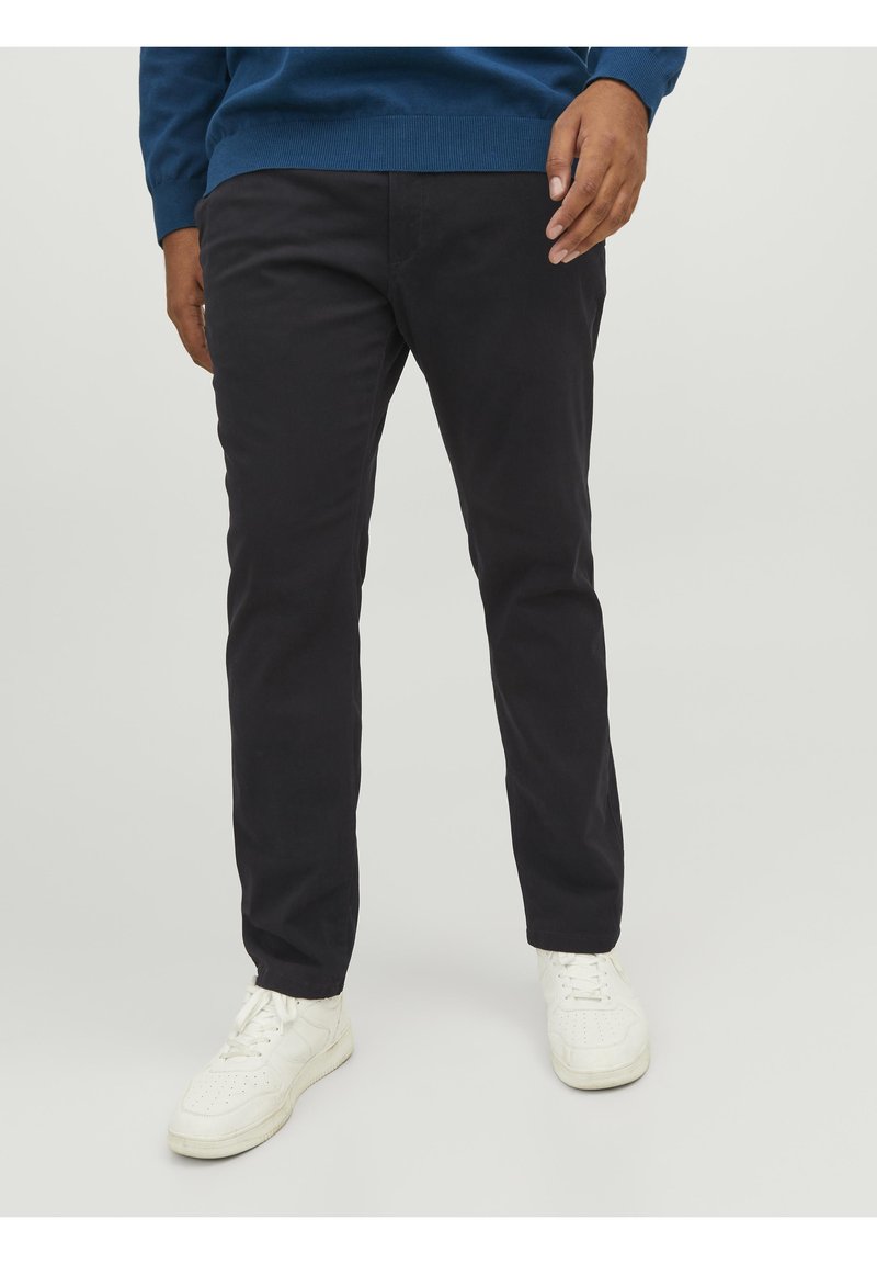 Homme portant un pantalon droit noir, des baskets blanches et un pull bleu, debout sur un sol et un arrière-plan gris clair.
