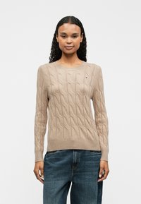Pull beige en maille torsadée avec des manches longues et un ourlet côtelé. Il présente un petit logo sur la poitrine, une texture douce et un col rond.