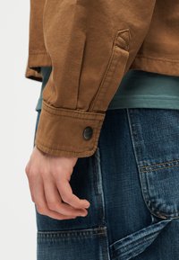 Hand ruht neben blauer Jeans mit großen Taschen, trägt eine braune Jackenärmel mit Umschlag und einem schwarzen Knopfdetail.