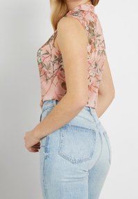 Blusa sin mangas floral en rosa con detalles en verde y marrón, combinada con vaqueros de talle alto en azul claro. Tela suave, ajuste relajado.