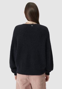 Vrouw met lang bruin haar, gekleed in een zwarte fuzzy trui en lichtroze jeans, van achteraf te zien tegen een grijze achtergrond.