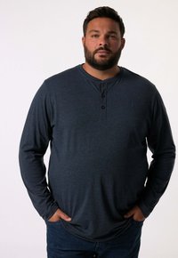 Tmavě modrá dlouhá rukávová košile typu henley s légou a třemi knoflíky, vyrobená z měkké tkaniny. Nositelná s modrými džíny, ruce položené v kapsách.