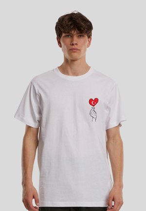 Ung mand med kort krøllet hår iført en hvid t-shirt med et motiv af en hånd, der holder et rødt hjerte med en halvmåne og stjerne symbol.